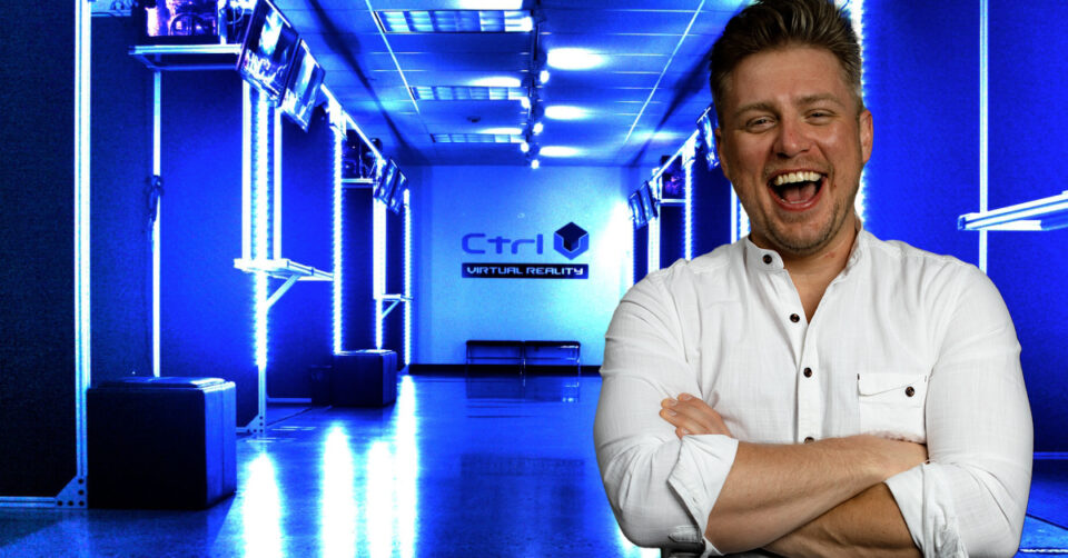 VR franchise CTrl V's CEO Robert Bruski stands, smiling, arms crossed, in a white button down shirt. He appears in front of a Ctrl V location interior.